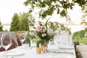 Wedding table setting decorated with fresh flowers in a brass vase. Wedding floristry. Banquet table for guests outdoors with a view of green nature. Bouquet with roses, eustoma and eucalyptus leaves
