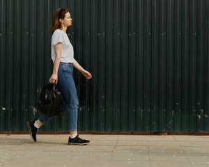 Female walking along fence. Side view of teenager wearing jeans and t-shirt and holding backpack...