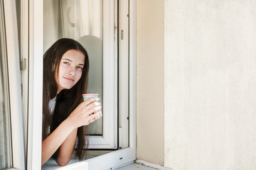 Fototapeta na wymiar young brunette hipster girl is looking out of open window of house, holding a colored paper disposable cup with coffee or tea in her hands, using disposable eco-friendly tableware