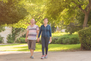 Obraz premium Family, age, sport, healthy and people concept - happy senior couple holding hands and exercising together. Active elderly man and woman fast walking outdoors in summer or autumn park on a sunny day.