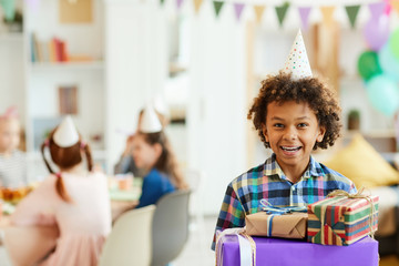 Portrait of happy African-American boy holding gift boxes posing during birthday party with friends, copy space - obrazy, fototapety, plakaty