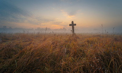 Lonely ancient tombstone in form of cross among high yellow grass at the autumn foggy field at dawn.