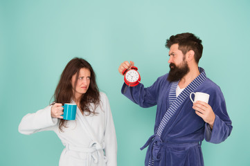 Time to wake up and have nice day. Guy husband hold coffee and clock. Every morning begins with coffee. Couple in bathrobes with mugs. Man with beard and sleepy woman enjoy morning coffee or tea