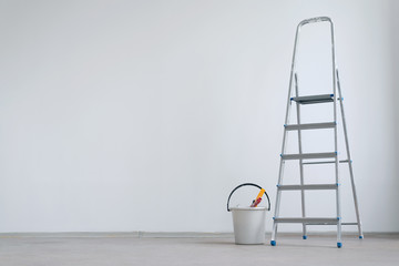 Ladder and bucket with paint roller on a white wall background with copy space. Under construction concept background. - obrazy, fototapety, plakaty