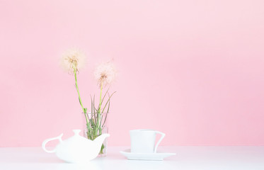 White porcelain teapot and cup on pink background with white dandelions. Concept for festive background.