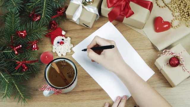 Female hand writing a wish list to Santa on wooden table with gifts, wrapping paper, red Christmas decorations. Xmas and Happy New Year composition. Flat lay, top view