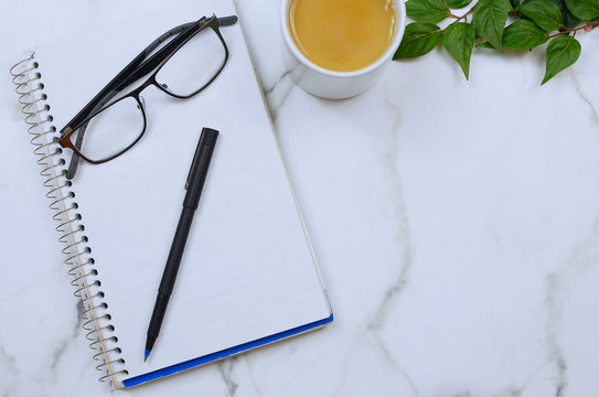 Flat lay on marble table top. Copy space. Items on table are business or school or office oriented and include a spiral notebook, a pen, reading glasses and a cup of coffee.