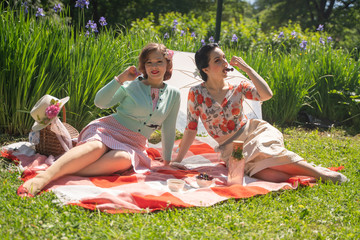 two pretty pin up ladies having nice picnic in the city park in a sunny day together. girls friends enjoy hot summer weather. beautiful females in retro vintage style are relaxing on the grass.