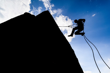 A dramatic silhouette of a climber rappeling down a rock wall. Rock climber with a rope abseil down. Mountaineer jumping down on climbing rope. Extreme adventure sport of rock climbing.