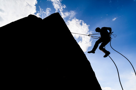 A dramatic silhouette of a climber rappeling down a rock wall. Rock climber with a rope abseil down. Mountaineer jumping down on climbing rope. Extreme adventure sport of rock climbing.