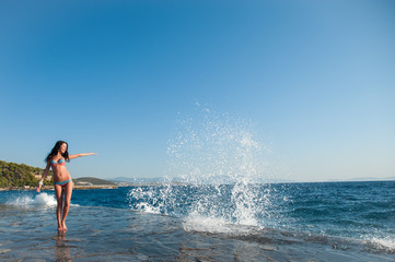 Fototapeta premium A girl in a swimsuit, splashing from the waves, Brunette girl rejoices splashing from the waves. Young happy woman jumping with raised arms, waving a blue bikini swimsuit in a blue swimsuit and a
