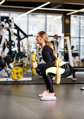 Fototapeta premium Slender girl with long blond hair dressed in sportswear is doing squats with plate in the modern gym