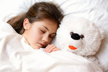 Small lovely girl with long dark hair healthy skin lies on comfortable bed with her favourite toy. Little girl sleeping with her teddy bear under the white warm blanket has pleasant dreams at night