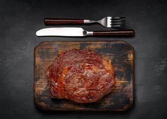 Ready to eat juicy grilled Rib Eye beef steak with crispy crust on wooden with knife and fork