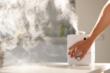 Hand turn on aroma oil diffuser on the table at home, steam from the air humidifier. Ultrasonic technology, increase in air humidity indoors, comfortable living conditions. 
