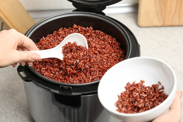 Woman putting brown rice into bowl from multi cooker in kitchen, closeup