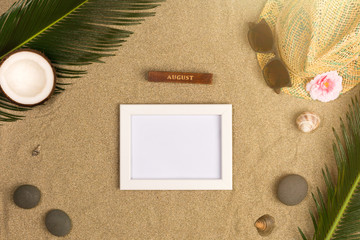 Fototapeta na wymiar Stylish summer composition with photo frame, green leaves, hat and sunglasses on a sand background. Artwork mockup with copy space