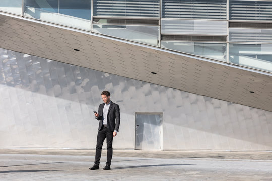 Businessman Using Mobile Phone App Texting Outside Of Office In Urban Modern City Building Background. Young Caucasian Man Holding Smartphone For Business Work.