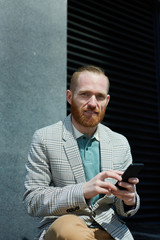 Smiling red-bearded manager in stylish jacket sitting outdoors and texting sms on smartphone while looking at camera