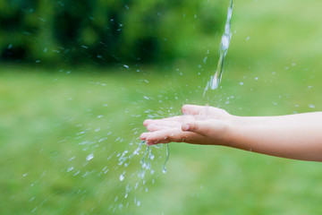 happy little girl catches a jet of water with her hands. child washing hand outdoors. Water pouring in kid hand on nature background, environment issues