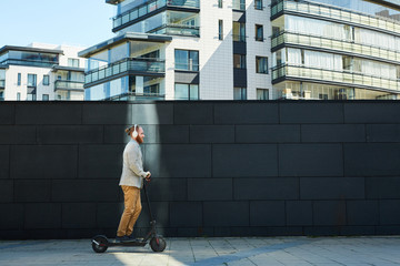 Side view of content sporty hipster businessman in headphones riding kick scooter over city street