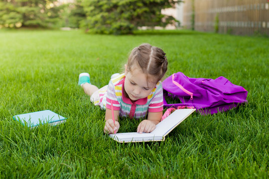 child girl schoolgirl elementary school student  lying on the grass and draws in a notebook. concept back to school. outdoor activities