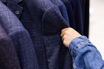 Rack With Suit Jackets In Department Store Or Clothes Boutique. Female Hand In Denim Jacket Checks Or Touches Fabric Of Men's Wool Jacket.