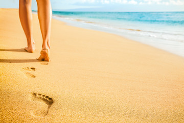 Beach woman legs feet walking barefoot on sand leaving footprints on golden sand in sunset. Vacation travel freedom people relaxing in summer.