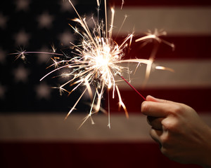 Hand holding lit sparkler in front of the American Flag for 4th of July celebration