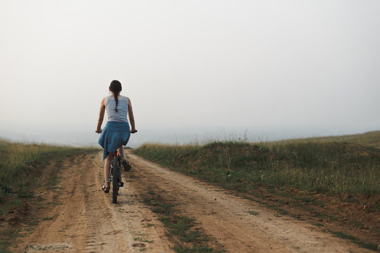 white caucasian young woman in casual clothing riding on bicycle in countryside road evening at sunset, view from back in full body size, lifestyles stock photo image