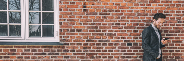 Happy business young man using mobile cellphone against city home brick wall background banner panorama.