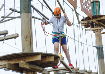 Boy in forest adventure park. Kid in orange helmet and white t shirt climbs on high rope trail. Climbing outdoor, amusement center for children. Young boy plays outdoors	