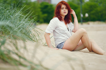 A young redheaded girl in a white shirt and jeans shorts sitting on the sand on the beach