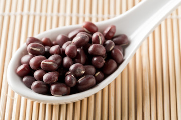 Raw, uncooked, dried adzuki (red mung) beans in white spoon on bamboo mat background