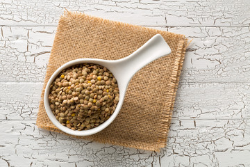 Raw, dry, uncooked brown lentil legumes in white bowl on wood table background top view flat lay...