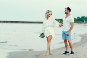 elegant and stylish woman with light curly hair dressed in a white blouse walking on the beach near water along with her elegant man in a white t-shirt and in a  blue shorts