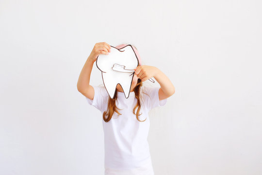 Baby girl kid holding paper image of big white tooth and toothbrush on grey background. Concept of health.