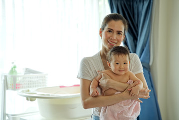 Asian beautiful mother holding little cute baby after taking a bath and dry her kid with towel standing in the room. Bathtub and bath shampoo are put in the background. Clean and healthy concept.