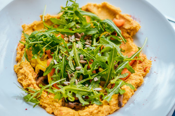 Bowl of carrot vegan hummus with chickpea, caramelized carrot, mashrooms, garlic, paprika, tahena, cumin, rocket salad and baked cauliflower.