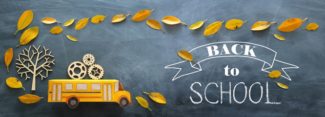 Fototapeta na wymiar Back to school. Top view banner of cardboard school bus with wooden gears as concept of success and achievement next to autumn dry leaves over classroom blackboard background