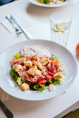 Plate of salad with tomatoes, jamon and baked vegetables on the white table. Glass of iced water with mint and lemon.