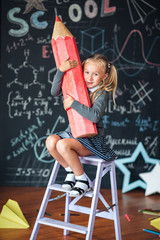 Back to school! A little happy blonde girl in school uniform sits with very large red pencil in her hands at school against chalkboard with school formulas . School Girl responds to the lesson.
