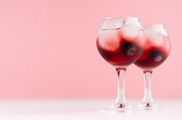 Exquisite arctic red alcoholic liquors with ice cubes, blueberry in two wineglasses on white wood table and pastel pink wall, copy space.