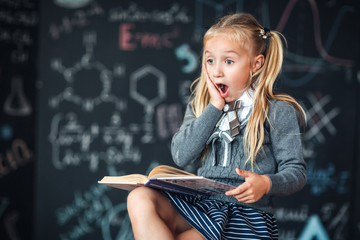 A sweet girl schoolgirl sits on a chalkboard with school formulas background. Works homework by reading a book Amazed to put his hands on his head, opening his mouth wide.