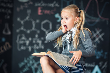 A sweet girl schoolgirl sits on a chalkboard with school formulas background. Works homework by reading a book Amazed to put his hands on his head, opening his mouth wide.