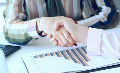 Friendly businesswoman handshaking over the office desk with economic charts after pleasant talk and effective negotiation, good relationships.