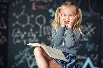 A sweet blond girl schoolgirl sits on a chalkboard with school formulas background. Works homework by reading a book Amazed to put his hands on his head, opening his mouth wide.