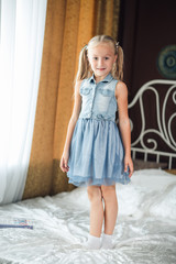 Cheerful girl standing on the bed. The girl laughs and jumps. On a white bed, the girl is having fun and jumping. Little baby in denim sundress jumping on white bed.