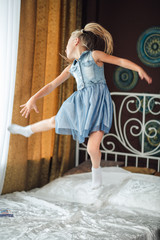 Cheerful girl jumping on the bed. The girl laughs and jumps. On a white bed, the girl is having fun and jumping. Little baby in denim sundress jumping on white bed.
