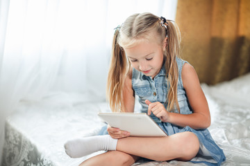 Cute little child girl blond in denim sundress lies in bed uses digital tablet. child playing on tablet pc.